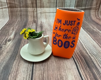 Puede incluir: Enfriador de latas naranja con el texto "I'm just here for the boos" en morado. El enfriador de latas tiene una telaraña, una araña y murciélagos impresos en él.
