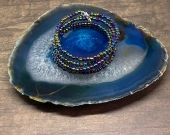 Puede incluir: Un brazalete de cuentas multicadena con cuentas iridiscentes azules, verdes y moradas. El brazalete está envuelto sobre sí mismo y está sentado sobre una rebanada de ágata azul y blanca.
