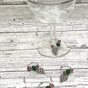 Puede incluir: Tres encantos para copa de vino de plata con cuentas verdes y moradas y un encanto de racimo de uva de plata. Los encantos están sobre una superficie de madera blanca.