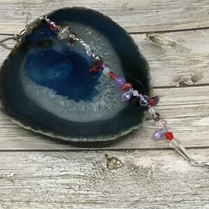 Op de afbeelding: Een kristallen zonenvanger met een zilveren haak en een verscheidenheid aan gekleurde kralen, waaronder rood, paars en roze. De zonenvanger hangt aan een blauwe en witte agaatplakje.