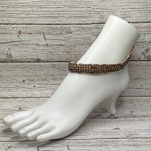 Op de afbeelding: Een gouden en bruine kralen enkelbandje op een witte mannequin voet.