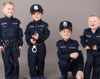May include: Four children wearing navy blue police uniforms with long-sleeved button-down shirts and matching trousers. Each shirt features the word POLICE and a name tag. The outfits include police caps, black belts, toy handcuffs, and flashlights.