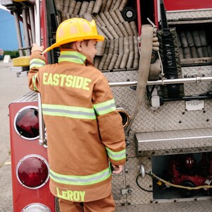 May include: A young person wearing a brown fire fighter costume with the words "Eastside" and "Leroy" on the back. The person is standing next to a red fire truck.