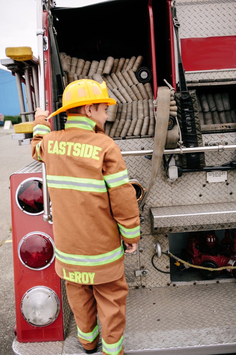 May include: A young person wearing a brown fire fighter costume with the words "Eastside" and "Leroy" on the back. The person is standing next to a red fire truck.