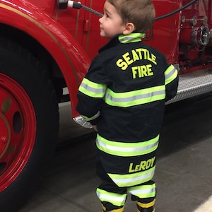 May include: A young child wearing a black firefighter uniform with yellow reflective stripes. The uniform has the words "Seattle Fire" and "Leroy" on it. The child is standing in front of a red fire truck.