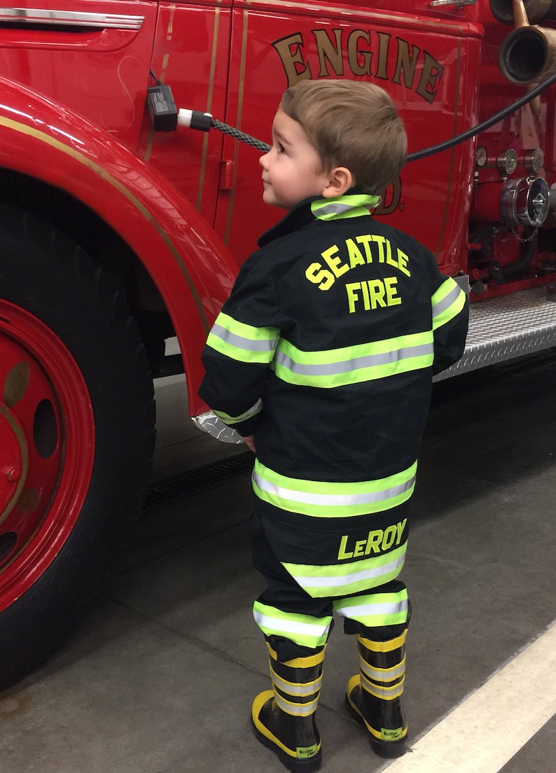 May include: A young child wearing a black firefighter uniform with yellow reflective stripes. The uniform has the words "Seattle Fire" and "Leroy" on it. The child is standing in front of a red fire truck.