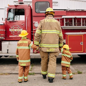 May include: A firefighter in a brown and yellow uniform with the name "Leroy" on the back stands in front of a red fire truck with two children in similar uniforms. The children are also wearing yellow helmets with the name "Leroy" on the front and "Eastside" on the back.