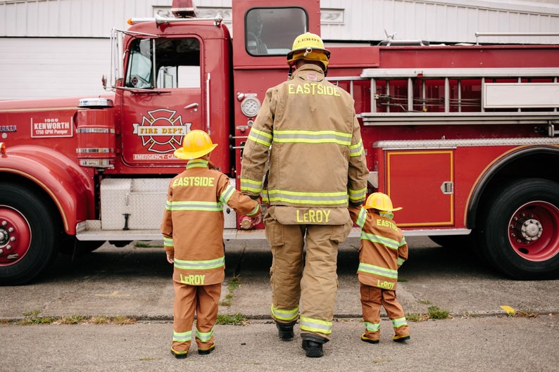 May include: A firefighter in a brown and yellow uniform with the name "Leroy" on the back stands in front of a red fire truck with two children in similar uniforms. The children are also wearing yellow helmets with the name "Leroy" on the front and "Eastside" on the back.