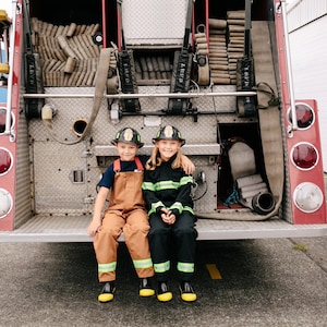 May include: Two children dressed as firefighters are sitting in the back of a red fire truck. They are wearing black and yellow fire suits and helmets. The fire truck has a ladder and hoses in the back.