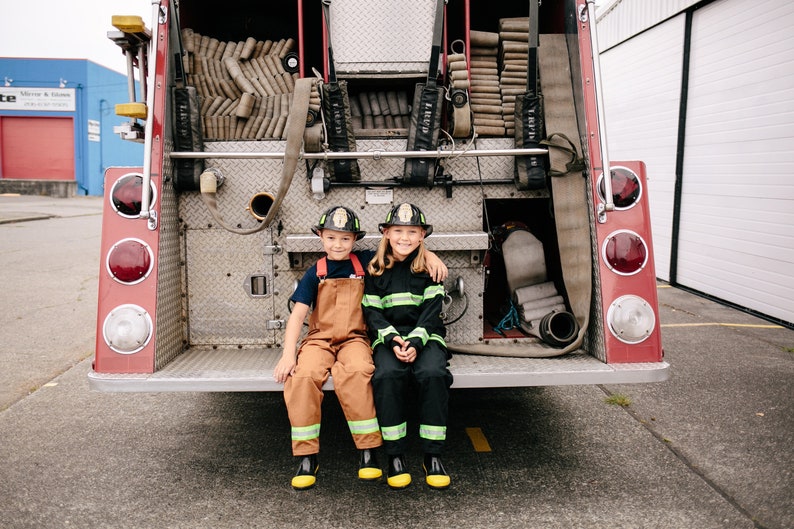 May include: Two children dressed as firefighters are sitting in the back of a red fire truck. They are wearing black and yellow fire suits and helmets. The fire truck has a ladder and hoses in the back.