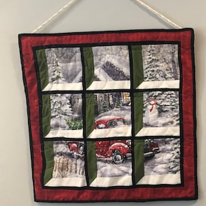 May include: A red and green quilted wall hanging with a window design. The window panes show a snowy winter scene with a red truck, a snowman, and a house.