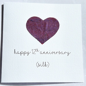 May include: A white card with a textured surface features a deep purple heart shape. The text "happy 12th anniversary (silk)" is written in a handwritten style. The card is a romantic gift.