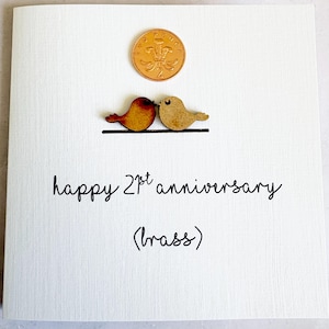 May include: A white greeting card featuring a copper-coloured two-pence coin above two small wooden bird cutouts perched on a black line. The text reads "happy 21st anniversary (brass)" in black script. The card has a subtle vertical texture.