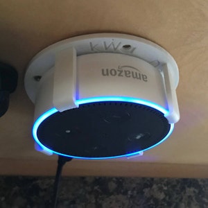 May include: White wall mount for an Amazon Echo Dot smart speaker. The speaker is black and has a blue light ring around the top. The mount has the word "amazon" printed on it.