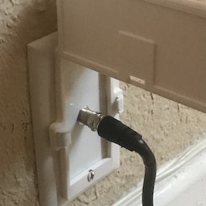 May include: A white wall plate with a coaxial cable connector and a black cable attached. The plate is mounted on a wall.