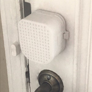 May include: A white door handle safety lock with a white plastic cover and a grid pattern. The lock is attached to a white door with a brass door handle.