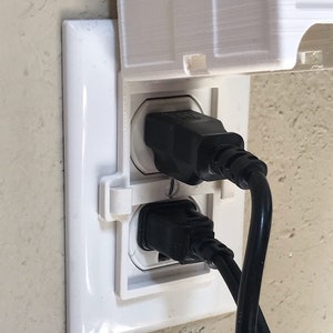 May include: A white electrical outlet with a white plastic cover that is partially open. Two black electrical cords are plugged into the outlet.