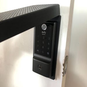 May include: Black smart lock with a keypad, fingerprint reader, and the "eufy SECURITY" logo. The lock is mounted on a white door, with a black handle visible in the upper left corner.