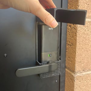 May include: Black smart lock with a lever handle and the brand name "eufy" visible. A hand is holding a black rectangular piece above the lock. The lock is mounted on a black door next to a brick wall.