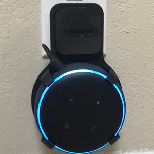 Peut inclure: Un haut-parleur intelligent Amazon Echo Dot de 4e génération noir avec un anneau lumineux bleu, monté sur un mur avec un support mural noir. Le haut-parleur est branché sur une prise électrique standard.