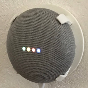 Puede incluir: Un altavoz inteligente Google Home gris con un soporte de pared blanco. El altavoz tiene una cubierta de tela y una fila de luces de colores.