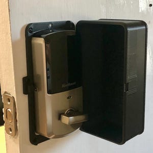 May include: A smart lock with a black and silver finish is mounted on a white door. The lock has a rectangular black cover and a silver handle. The brand name "Kwikset" is visible on the lock.