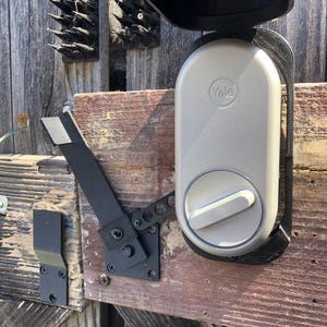 Könnte beinhalten: Ein hellgraues Yale Smart Lock ist an einem verwitterten Holztor montiert. Das Schloss hat ein kreisförmiges Zifferblatt und einen schwarzen Riemen. Ein schwarzer Metallverschluss und Scharniere sind ebenfalls sichtbar.