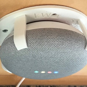 May include: White wall mount for a Google Home smart speaker. The mount has two white plastic arms that hold the speaker in place. The speaker is gray with a fabric mesh covering and has a white plastic ring around the top. The speaker has five colored lights on the front.