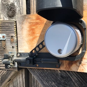 May include: A silver smart lock with a black protective cover, mounted on a wooden door frame. The lock has a round face with a small logo and a black lever.