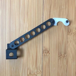 May include: A black and white bottle opener with a distinctive design. It has a long, slotted arm attached to a square base and a curved, bottle-cap-shaped end. The opener is secured with small screws and sits on a wooden surface.