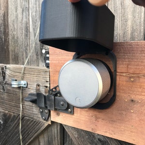 May include: A silver smart lock with a black protective cover, mounted on a wooden door. The lock has a circular design with a small logo in the centre.