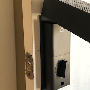 Op de afbeelding: Zwart slim slot met de merknaam "eufy SECURITY" op een witte deur. Het slot heeft een zwarte knop en een rechthoekig ontwerp. Een zwart rechthoekig apparaat is boven het slot gemonteerd.