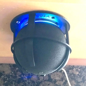 Pode incluir: Um alto-falante preto com um anel de luz azul está montado no teto com uma gaiola preta impressa em 3D. A gaiola tem a palavra "USA" impressa nela.