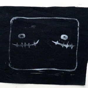 May include: A black fabric square with a white, hand-drawn design. The design features two stylized faces within a rectangular border. Each face has a circular eye and a jagged, tooth-like mouth. The artwork is simple and graphic.