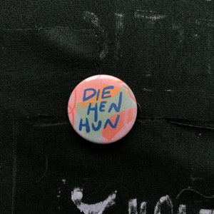 Puede incluir: Un bot&oacute;n redondo con el texto "DIE HEN HUN" en azul, naranja y verde. El bot&oacute;n tiene un fondo rosa y blanco con trazos de pintura abstractos. El bot&oacute;n est&aacute; sobre una superficie de tela negra.