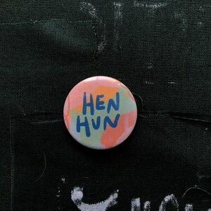 Puede incluir: Un bot&oacute;n redondo con el texto "HEN HUN" en azul, sobre un fondo de colores pastel que incluye rosa, naranja y verde. El bot&oacute;n est&aacute; sobre una superficie de tela negra.