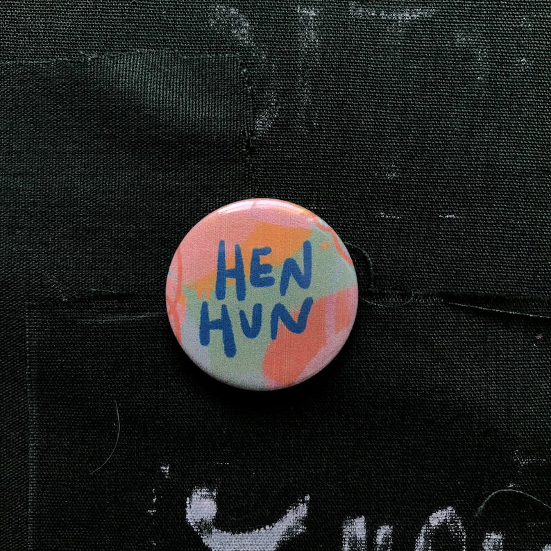 Puede incluir: Un bot&oacute;n redondo con el texto "HEN HUN" en azul, sobre un fondo de colores pastel que incluye rosa, naranja y verde. El bot&oacute;n est&aacute; sobre una superficie de tela negra.