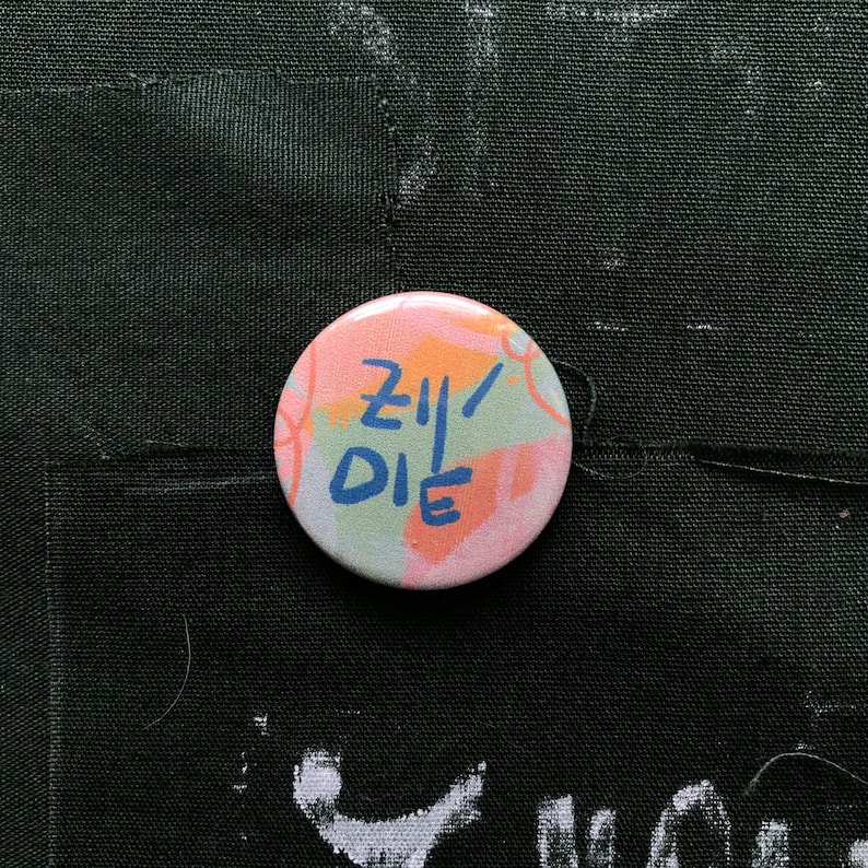 Puede incluir: Un bot&oacute;n redondo con una combinaci&oacute;n de colores pastel rosa, naranja y verde. El bot&oacute;n presenta el texto azul "ZII DIE" en un estilo manuscrito. El bot&oacute;n est&aacute; sobre un fondo de tela verde oscuro.