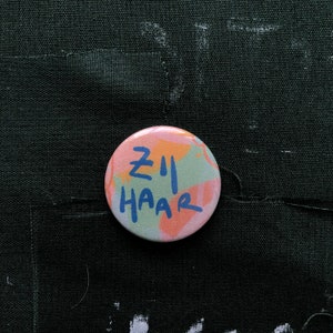 Puede incluir: Un bot&oacute;n redondo con el texto "ZII HAAR" en azul, sobre un fondo colorido de pinceladas rosas, verdes y naranjas. El bot&oacute;n est&aacute; colocado sobre una tela verde oscuro.
