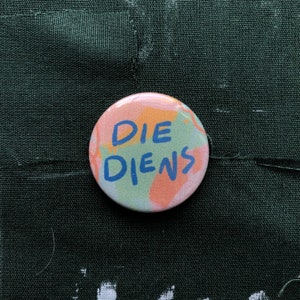 Puede incluir: Un bot&oacute;n redondo con las palabras "DIE DIENS" en letras azules. El bot&oacute;n tiene un fondo colorido con pinceladas rosas, naranjas y verdes. Se encuentra sobre un fondo de tela verde oscuro.