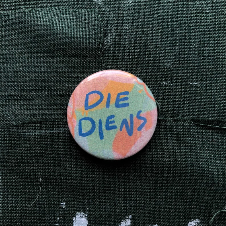 Puede incluir: Un bot&oacute;n redondo con las palabras "DIE DIENS" en letras azules. El bot&oacute;n tiene un fondo colorido con pinceladas rosas, naranjas y verdes. Se encuentra sobre un fondo de tela verde oscuro.