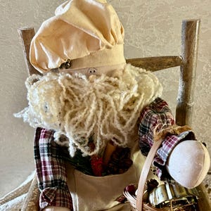 May include: A handcrafted Santa Claus doll, seated on a rustic wooden chair. The doll features a cream-colored chef's hat, a fluffy white beard, and a plaid shirt. It holds a small woven basket, and its outfit includes red and cream striped pants.