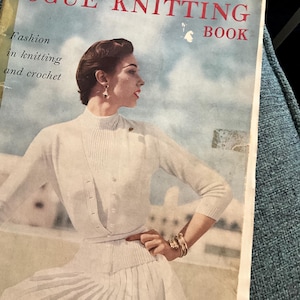 Puede incluir: Una portada de libro vintage de Vogue Knitting con una mujer que lleva un cárdigan y una falda de punto blanco. La portada es azul con nubes blancas y el texto "Vogue Knitting Book" en rojo.