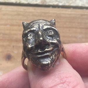 Puede incluir: Un anillo de bronce con una escultura detallada de la cara de un diablo. El anillo se lleva en un dedo.
