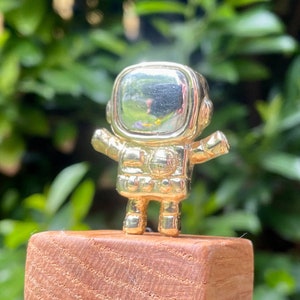 May include: A gold astronaut figurine standing on a wooden platform. The astronaut is wearing a helmet with a clear visor and has a small, round body.