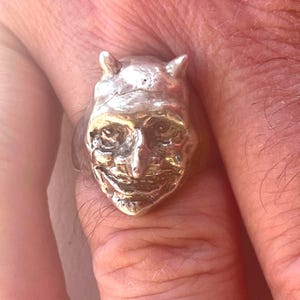 Peut inclure: Une bague argentée représentant un visage sculpté en trois dimensions avec des cornes pointues, des yeux enfoncés, un nez proéminent et une large bouche souriante. La surface métallique présente une finition polie et réfléchissante.