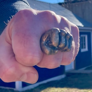 Pode incluir: Um close-up de uma mão usando um grande anel de prata em forma de punho fechado.