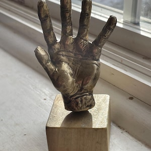 Könnte beinhalten: Eine bronzefarbene Skulptur einer erhobenen Hand, montiert auf einem quadratischen Holzsockel. Die Hand ist detailliert, und die Skulptur befindet sich in der Nähe eines Fensters.