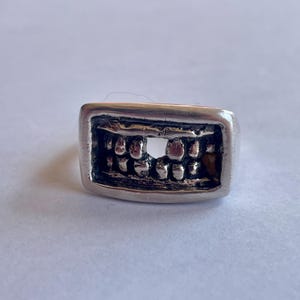 Può includere: Anello in argento con una superficie rettangolare che presenta un design dettagliato di una dentatura. I denti sono scolpiti individualmente, con uno spazio al centro. L'anello ha una finitura lucida e un'estetica audace e unica.