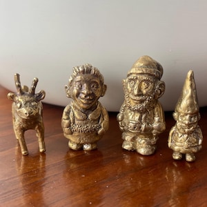 Puede incluir: Un conjunto de cuatro figuritas doradas, que incluyen un ciervo, una mujer, un hombre con barba y un gnomo. Las figuritas están todas de pie y tienen una calidad caprichosa y de cuento de hadas.